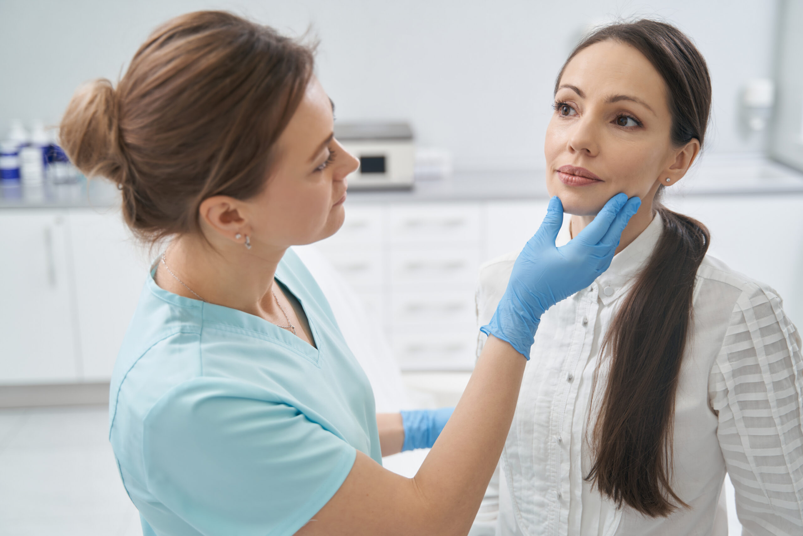 Medical professional in blue gloves examines a patient's face during a checkup in a bright clinic office.